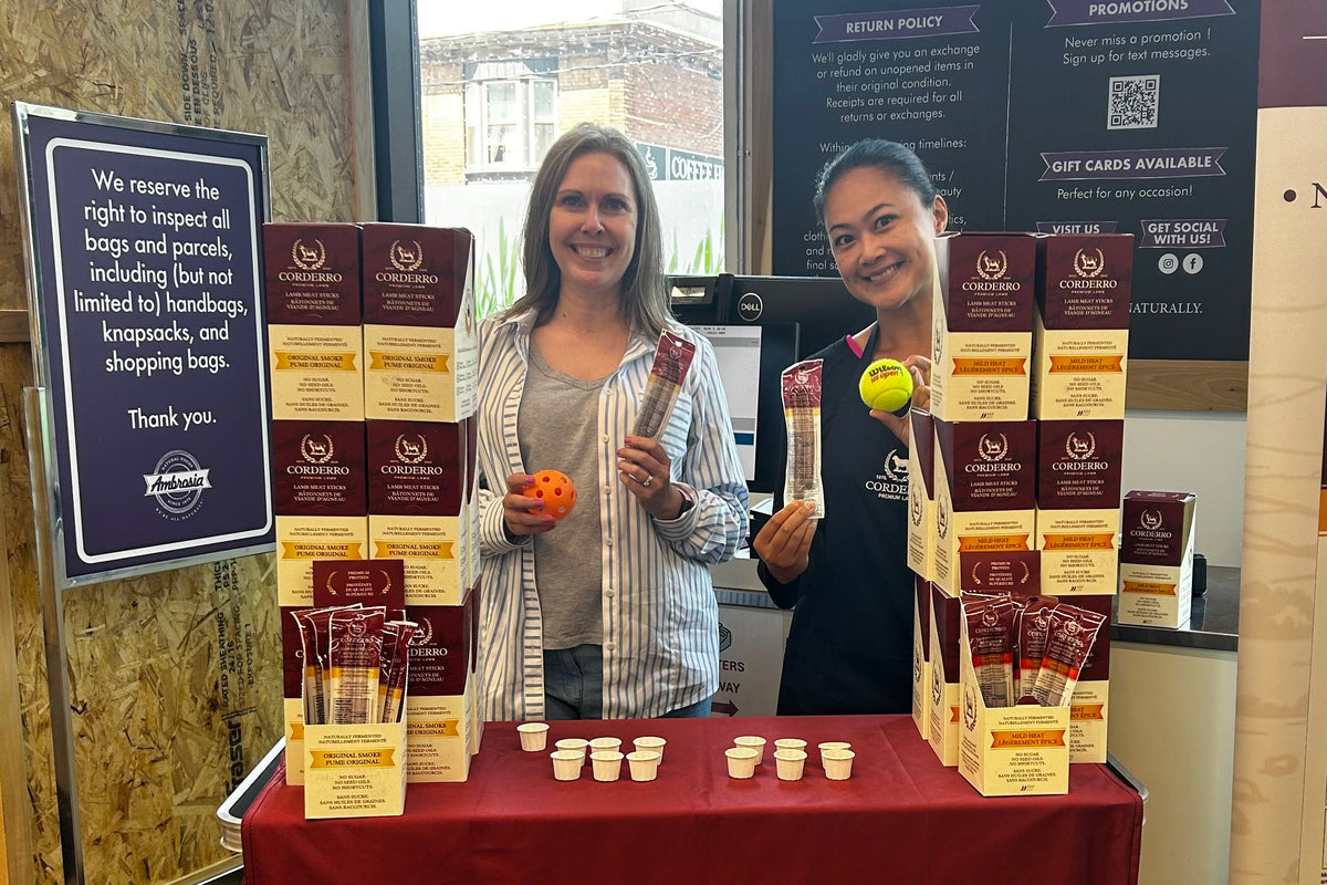 Two women stand behind a branded Corderro Premium Lamb display table at an indoor event. The table is covered with a deep red Corderro cloth and covered with multiple boxed and bagged lamb products, small cups, and two people holding packaged food items.