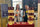 Two women stand behind a branded Corderro Premium Lamb display table at an indoor event. The table is covered with a deep red Corderro cloth and covered with multiple boxed and bagged lamb products, small cups, and two people holding packaged food items.